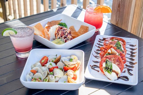 A table with three dishes: seafood salad, caprese salad, and a bowl of crispy snacks, alongside colorful drinks.