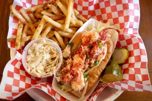 A lobster roll topped with lobster meat, served with fries, coleslaw, and pickles in a checkered basket.