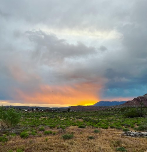 A scenic landscape featuring a colorful sunset behind mountains, with grassy fields and a cloudy sky.
