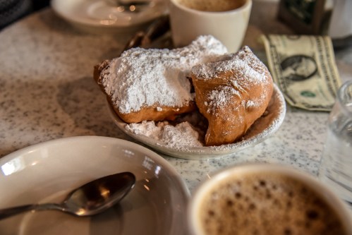 A plate of beignets dusted with powdered sugar, accompanied by coffee and a dollar bill on a table.