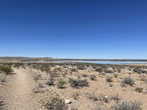 A clear blue sky over a desert landscape with sparse vegetation and a calm body of water in the distance.