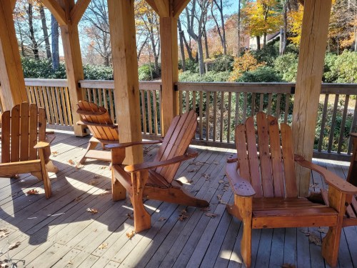 Four wooden Adirondack chairs on a porch surrounded by autumn foliage and trees.