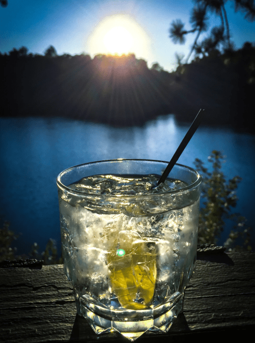 A glass of cocktail with ice sits on a railing, overlooking a serene lake at sunset.