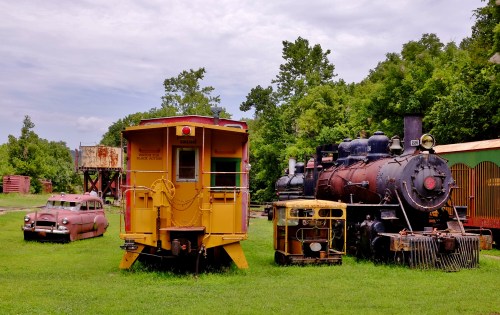 A vintage yellow train car and steam locomotive beside an old car, set in a grassy area with trees in the background.