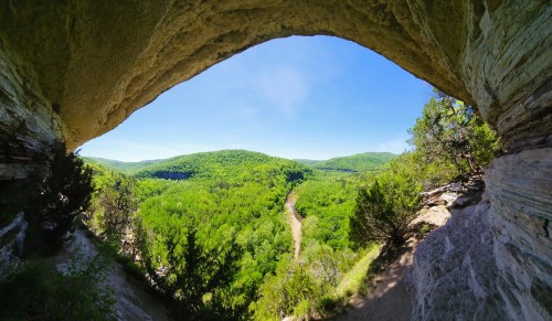A panoramic view from a rocky overhang, showcasing lush green hills and a winding river under a clear blue sky.