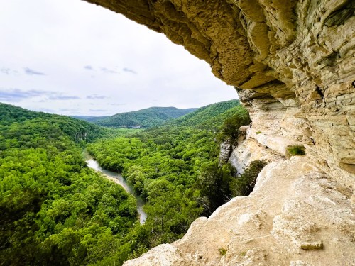 A scenic view from a rocky ledge overlooking a lush green valley and winding river below.