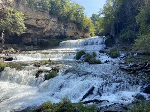 A serene waterfall cascading over rocky ledges, surrounded by lush greenery and a clear blue sky.