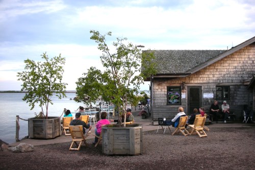A lakeside scene with people sitting in wooden chairs near a rustic building and trees, enjoying the view.