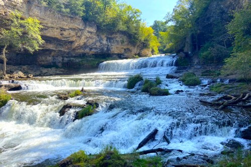 A serene waterfall cascades over rocky ledges, surrounded by lush greenery and sunlight filtering through the trees.