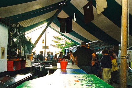 A bustling market scene under a large tent, with people mingling and colorful clothing hanging above.