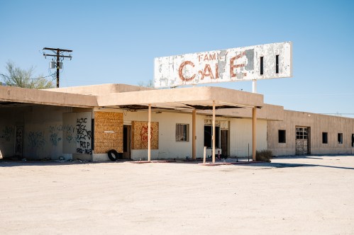 Abandoned café building with a faded sign, surrounded by desert landscape and graffiti on the walls.