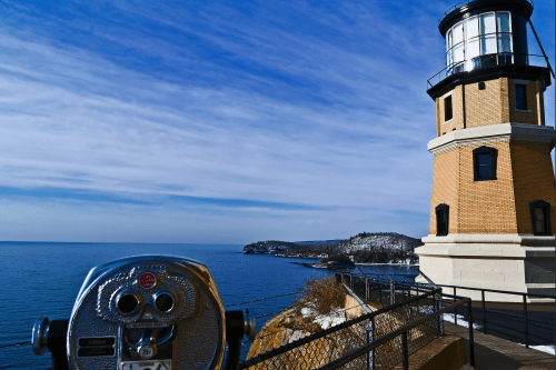 A scenic view of a lighthouse by the ocean, with a coin-operated viewer in the foreground and a clear blue sky.