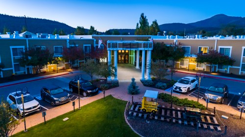 A hotel entrance at dusk, featuring columns, parked cars, and landscaped greenery with mountains in the background.