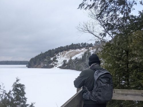 A person in a black beanie and jacket stands on a wooden overlook, gazing at a snowy lake and forested hillside.