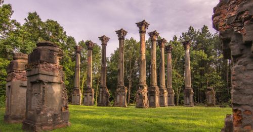 Ancient stone columns from the Windsor Ruins in MS stand amidst lush greenery, remnants of a historical structure under a cloudy sky.