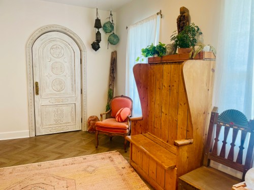 Cozy interior featuring a wooden bench, vintage chair, ornate door, and plants in a bright, inviting space.