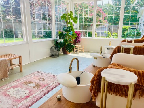 A bright sunroom with foot baths, plants, and a cozy rug, featuring large windows overlooking a garden.