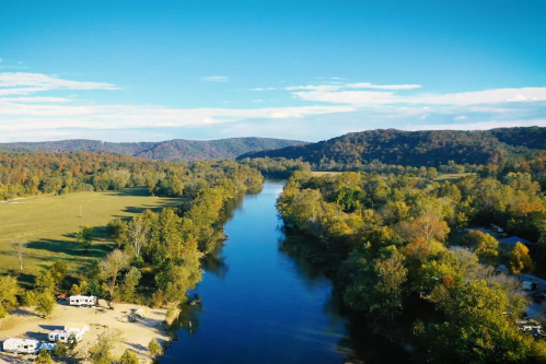Aerial view of a serene river surrounded by lush green trees and rolling hills under a clear blue sky.