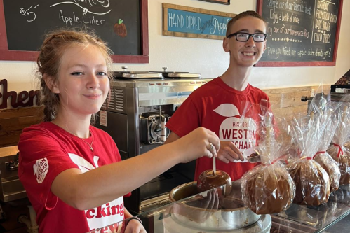 Two young people in red shirts prepare caramel apples at a counter, smiling in a cozy shop setting.