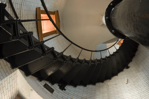 A spiral staircase viewed from below, featuring black metal steps and white tiled walls, with a window at the top.