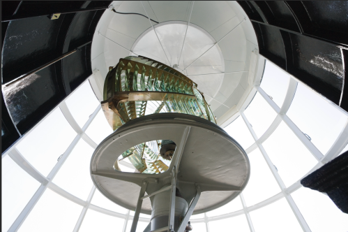 View from inside a lighthouse, looking up at the glass lens and circular structure against a bright sky.