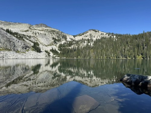 A serene lake reflecting mountains and trees under a clear blue sky, with a rocky shore in the foreground.