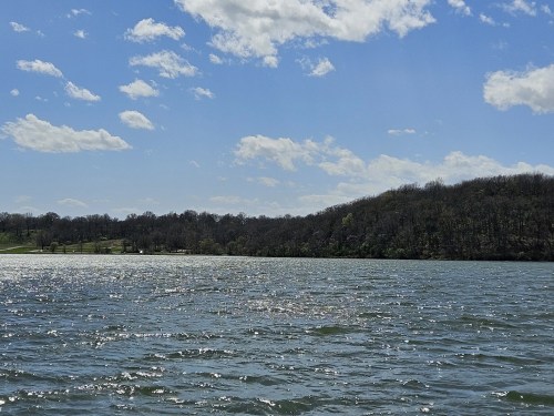 A serene lake under a blue sky with fluffy clouds, surrounded by lush green hills and shimmering water.