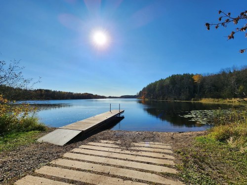 A serene lake scene with a wooden dock, clear blue sky, bright sun, and autumn foliage along the shoreline.