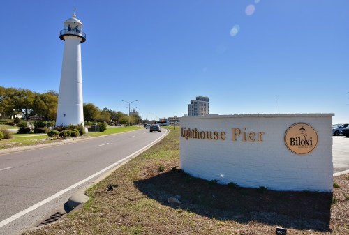 A white lighthouse stands beside a sign for Lighthouse Pier in Biloxi, with clear blue skies and a road in view.