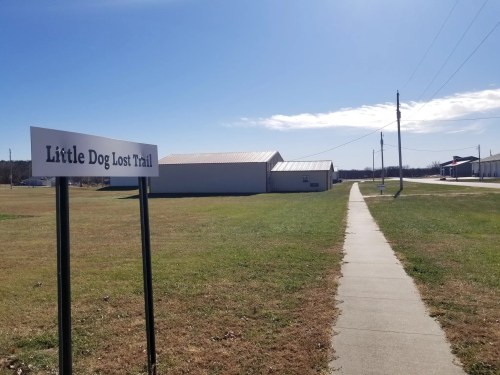 Sign for "Little Dog Lost Trail" next to a paved walkway, with buildings and a clear blue sky in the background.