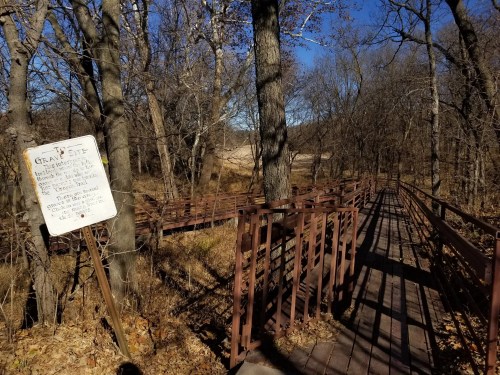 A wooden boardwalk through a wooded area, with a sign indicating a grave site nearby. Trees are bare in autumn.
