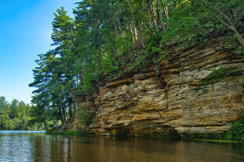 A serene river scene with a rocky cliff and lush green trees under a clear blue sky.