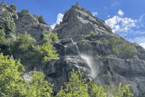A waterfall cascading down rocky cliffs, surrounded by lush greenery and a clear blue sky with scattered clouds.