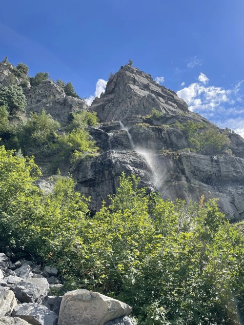A rocky mountain with a waterfall cascading down, surrounded by lush greenery and a clear blue sky.