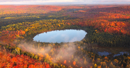 Aerial view of a vibrant autumn landscape with a circular lake surrounded by colorful fall foliage and mist.