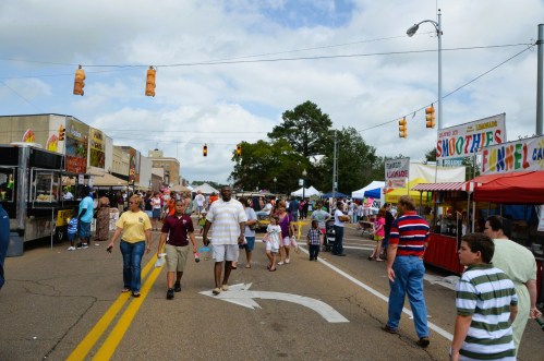 A busy street fair with people walking, food stalls, and colorful tents under a cloudy sky.