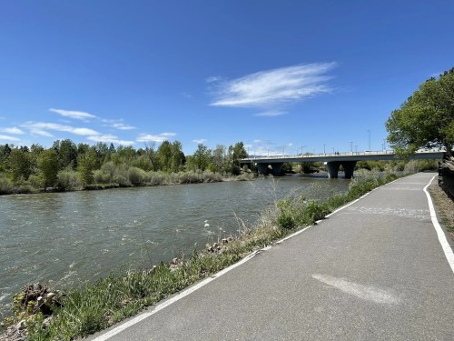 A scenic river view with a pathway alongside, under a clear blue sky and a bridge in the background.