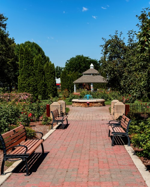 A serene park pathway lined with benches, leading to a gazebo surrounded by lush greenery and blooming flowers.