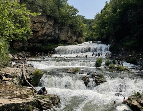 A scenic waterfall with multiple tiers, surrounded by lush greenery and people enjoying the water below.