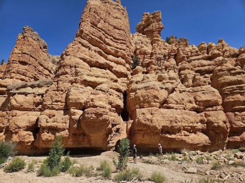 Two hikers explore a rugged, orange rock formation under a clear blue sky. Lush greenery is visible at the base.