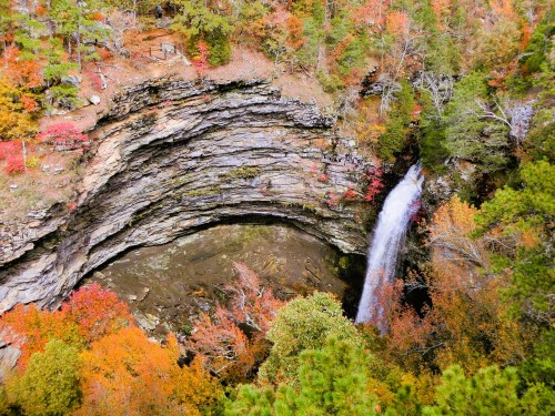Aerial view of a waterfall cascading into a rocky basin, surrounded by vibrant autumn foliage.