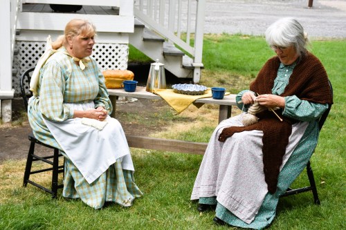 Two women in period clothing sit outdoors, one knitting and the other holding a piece of fabric, with a table set nearby.