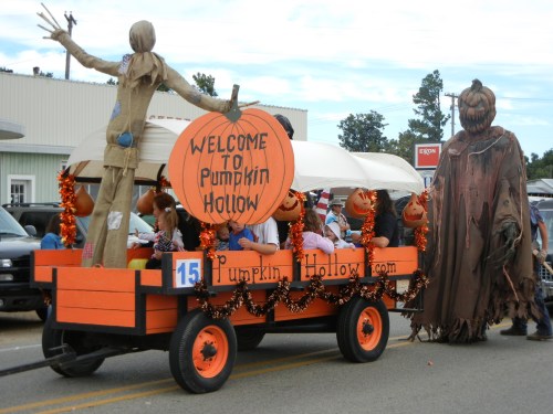 A festive parade float with pumpkins, a scarecrow, and a costumed figure, welcoming visitors to Pumpkin Hollow.
