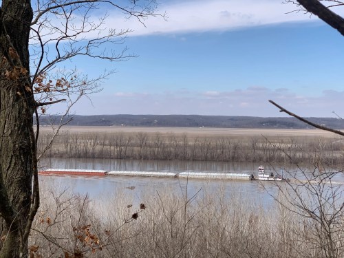 A river scene with a barge traveling along the water, framed by trees and a distant landscape under a blue sky.