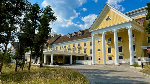 Historic yellow hotel with white columns, surrounded by trees and a blue sky, featuring a driveway and parked vehicle.