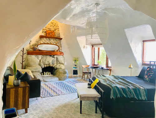 A bed is positioned by a large rock fireplace in a white room with plastered, hay bale walls.