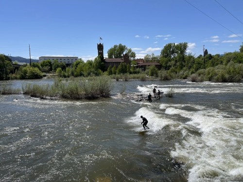 A river with small waves, featuring surfers and a scenic backdrop of trees and a building under a clear blue sky.
