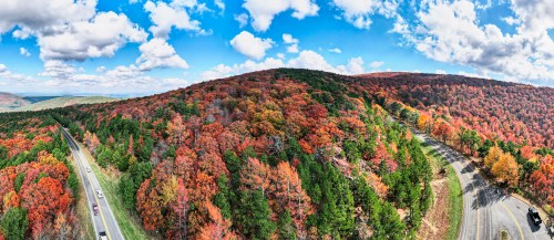 A panoramic view of vibrant autumn foliage on rolling hills, with a winding road and blue sky dotted with clouds.