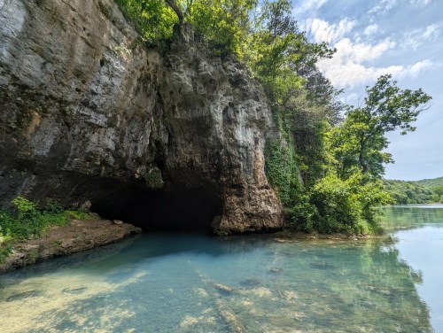 A serene river scene featuring a rocky cave entrance surrounded by lush greenery and clear blue water.