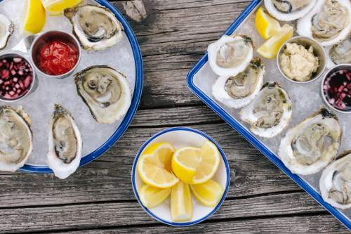 A platter of fresh oysters on ice, served with lemon wedges and various sauces in small bowls.